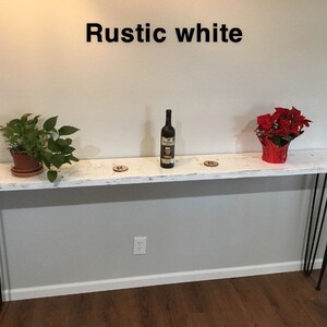 May include: White console table with black hairpin legs. The table is decorated with a bottle of wine, two potted plants, and a red poinsettia plant. The text "Rustic white" is written above the table.