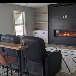 May include: A living room with a black fireplace and a long wooden table with hairpin legs. There are three black leather chairs and three bar stools in front of the table. The room has a white rug with black geometric patterns.
