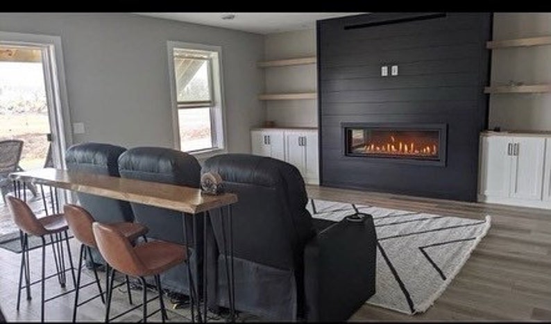 May include: A living room with a black fireplace and a long wooden table with hairpin legs. There are three black leather chairs and three bar stools in front of the table. The room has a white rug with black geometric patterns.