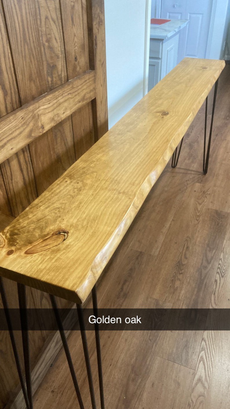 May include: A wooden console table with a live edge top and hairpin legs. The table is made of golden oak wood and has a natural finish. The table is sitting on a hardwood floor.
