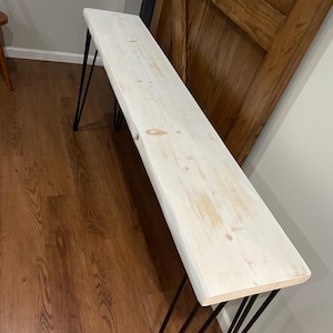 May include: Whitewashed wood console table with black hairpin legs. The table has a rustic, farmhouse style.