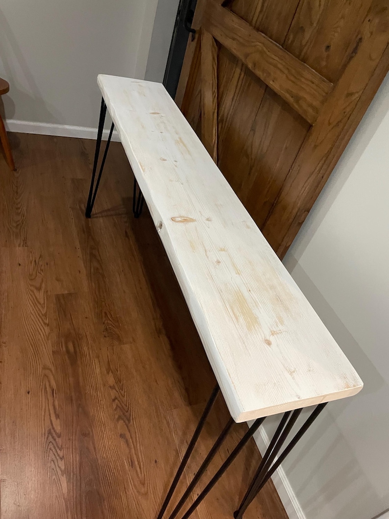 May include: Whitewashed wood console table with black hairpin legs. The table has a rustic, farmhouse style.