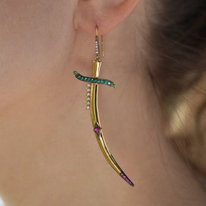 Könnte beinhalten: Goldene Ohrringe in Form eines gebogenen Schwertes mit grünen und rosafarbenen Edelsteinen.