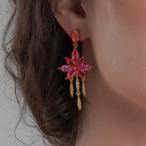Può includere: Orecchini pendenti dorati con un designo a forma di fiore rosa e dettagli in cristallo trasparente.