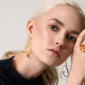 May include: A woman wearing gold geometric earrings and a gold ring with a circular design. The ring is on her right hand, and the earrings are on both ears.