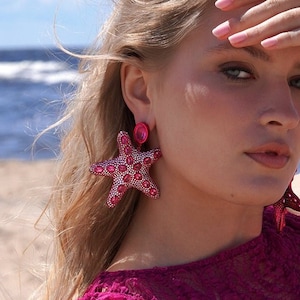 Puede incluir: Una mujer con un vestido de encaje rosa con un broche de estrella de mar rosa y pendientes de estrella de mar rosa. Está tumbada en una playa de arena.