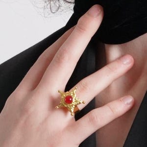 May include: A gold-toned ring featuring a five-pointed star design with a textured, hammered surface. The center of the star is set with a smooth, round, opaque red stone. The ring band is plain and metallic, encircling the finger.