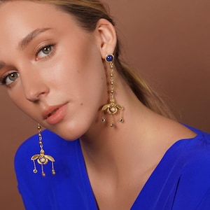Könnte beinhalten: Eine Frau in einem königsblauen Kleid mit goldenen Ohrringen, die mit einem blauen Edelstein und einem floralen Design verziert sind.