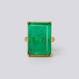 Op de afbeelding: Een gouden ring met een grote, rechthoekige, smaragdgeslepen groene edelsteen. De smaragd is gezet in een gouden bezel met een getextureerde band. De ring wordt getoond tegen een effen, lichte achtergrond.