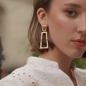 May include: A pair of gold tone earrings with a rectangular design. The earrings are made of a cream colored material and have red crystal accents.