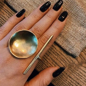 Può includere: Un anello color oro con un grande design rotondo a forma di ciotola e una lunga fascia dritta. L'anello è mostrato su una mano con smalto nero.
