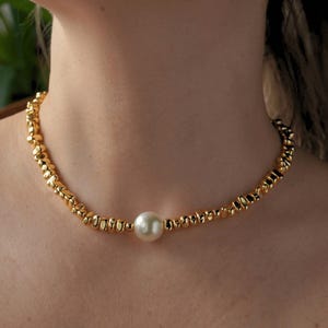 May include: A gold necklace with a large, round, white pearl pendant. The necklace is made of small, textured gold beads. The jewellery is displayed around a person's neck, highlighting the necklace's design and colour.