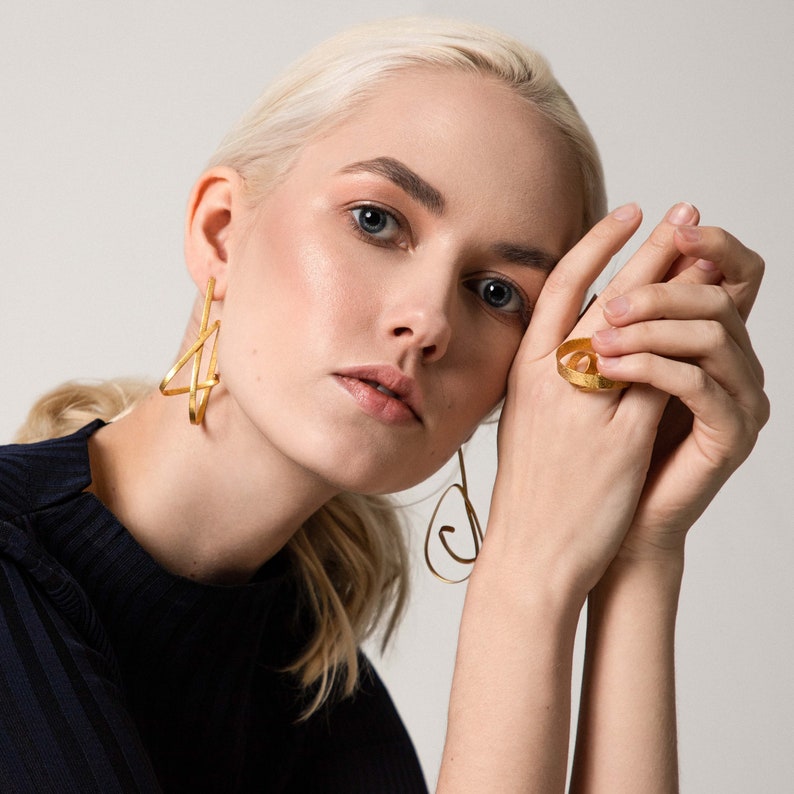 May include: A woman wearing gold geometric earrings and a gold ring with a circular design. The ring is on her right hand, and the earrings are on both ears.