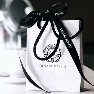 May include: White gift bag with black ribbon and a black and white logo that says "Dinari Jewels".