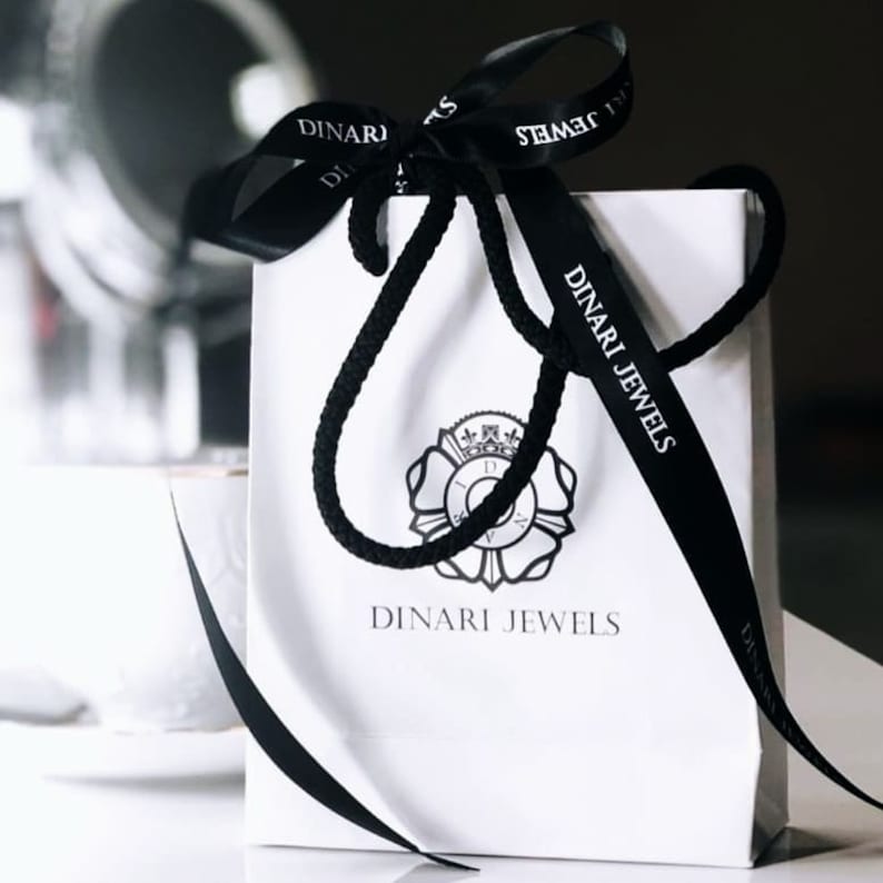 May include: White gift bag with black ribbon and a black and white logo that says "Dinari Jewels".