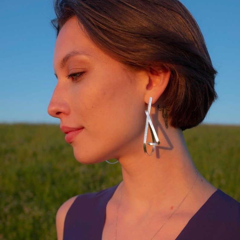 May include: A woman wearing silver geometric earrings. The earrings are made of thin metal strips that form a triangle shape. The woman is wearing a purple shirt and has short brown hair.