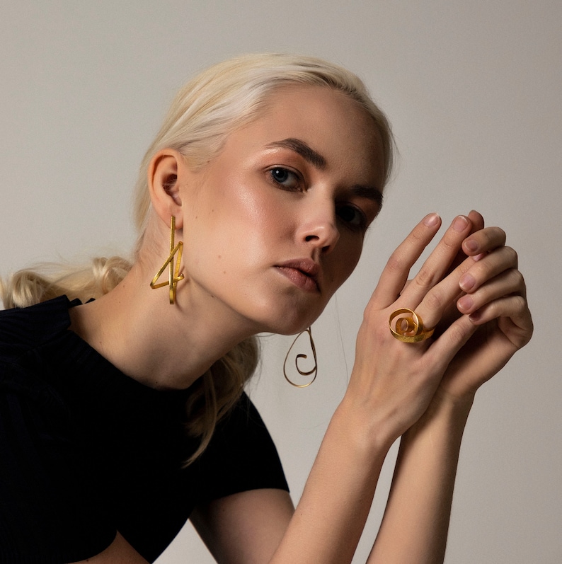 May include: A woman wearing gold geometric earrings and a gold ring with a spiral design. The ring is on her right hand, and the earrings are on both ears.