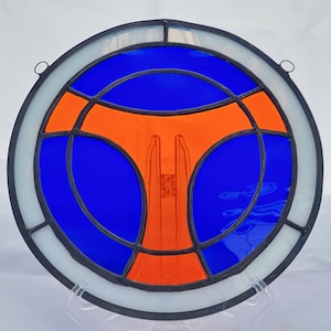 Puede incluir: Una ventana redonda de vitrales con un diseño geométrico azul y naranja. La ventana está enmarcada en metal negro y tiene un borde blanco.