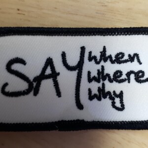 Puede incluir: Parche rectangular blanco con texto bordado en negro que dice "SAY when where why".