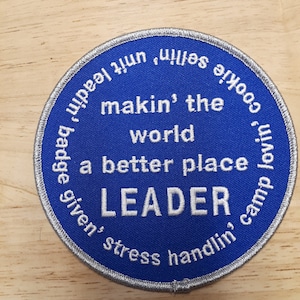 Peut inclure: Écusson rond bleu avec du texte blanc qui dit "Cookie lovin', camp given, stress handlin', makin' the world a better place LEADER.  Selfies within 2 min."