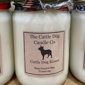 May include: Three glass jars with white candles and brown paper labels. The center jar has a red and black plaid ribbon and the text "The Cattle Dog Candle Co. Cattle Dog Kisses Hand Poured in Ohio 12 ounce soy".
