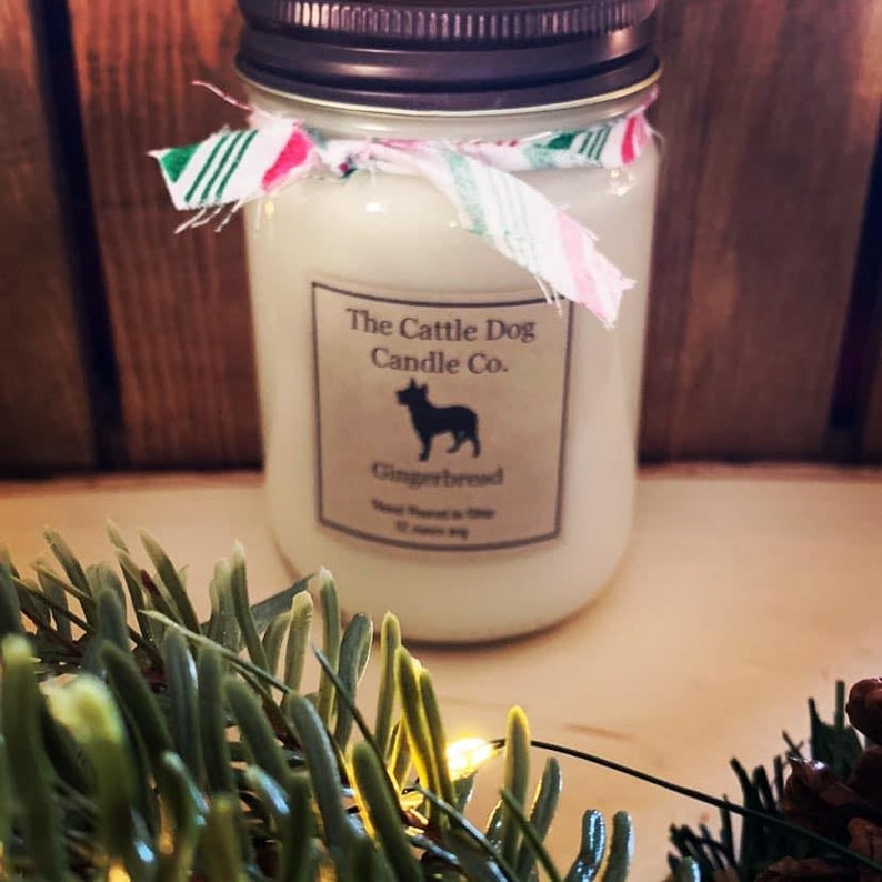 May include: A white candle in a glass jar with a brown lid. The jar is decorated with a white and green striped ribbon. The label on the jar reads "The Cattle Dog Candle Co." and "Gingerbread".