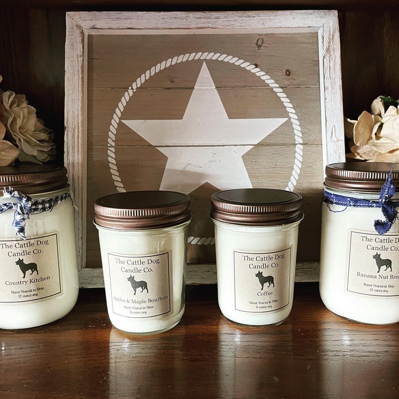 May include: Four glass candle jars with brown lids and white labels. The labels feature a black silhouette of a dog and text that reads "The Cattle Dog Candle Co." and the scent of each candle. The candles are arranged on a wooden surface in front of a wooden sign with a white star and rope design.