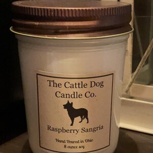 May include: A white candle jar with a brown lid. The label reads "The Cattle Dog Candle Co." and features a silhouette of a dog. The candle is labeled "Raspberry Sangria" and states "Hand Poured in Ohio 8 ounce soy".
