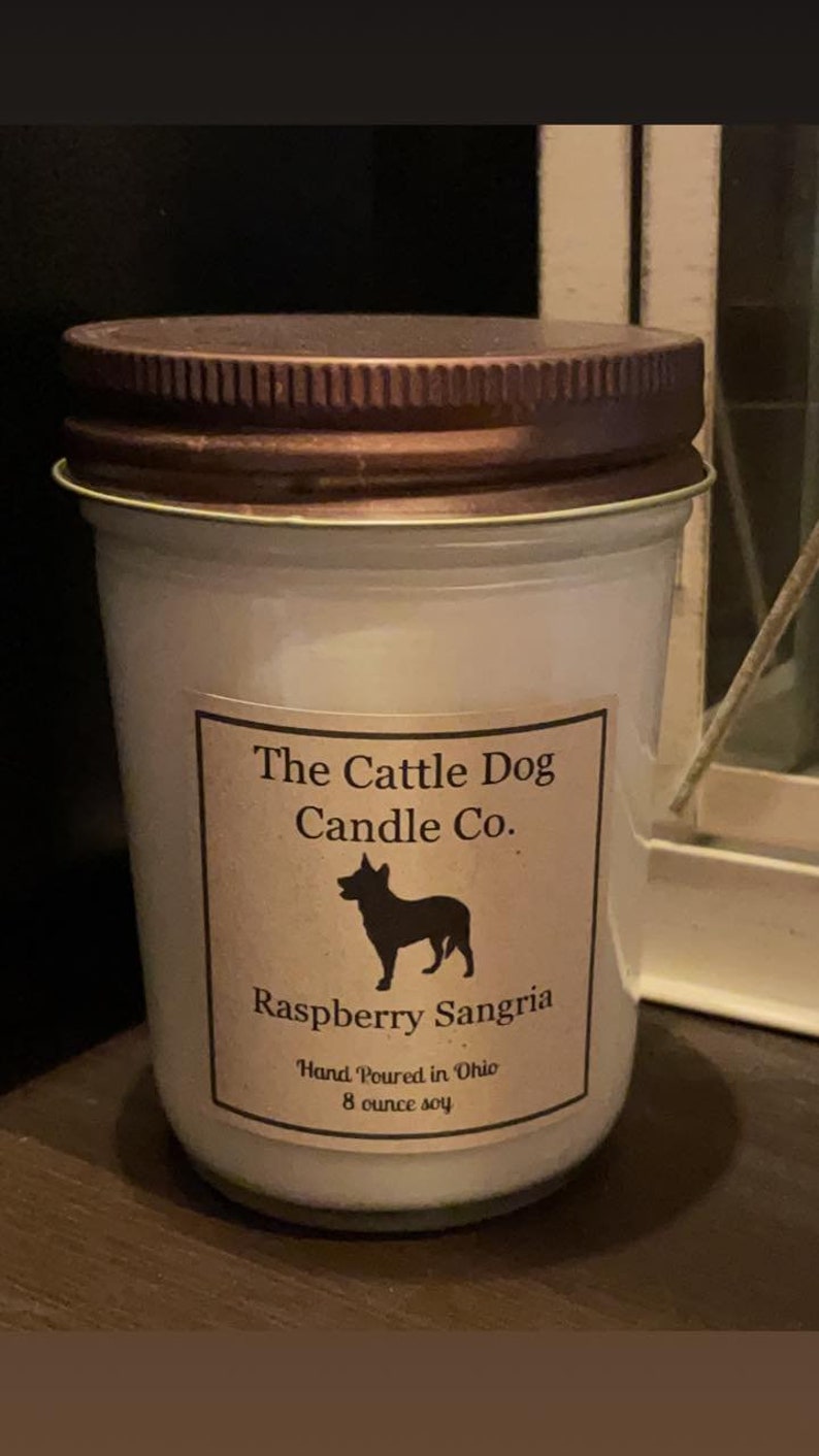 May include: A white candle jar with a brown lid. The label reads "The Cattle Dog Candle Co." and features a silhouette of a dog. The candle is labeled "Raspberry Sangria" and states "Hand Poured in Ohio 8 ounce soy".