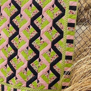 May include: A quilt with a geometric pattern of black, green, and pink squares. The quilt is quilted with a swirl design.