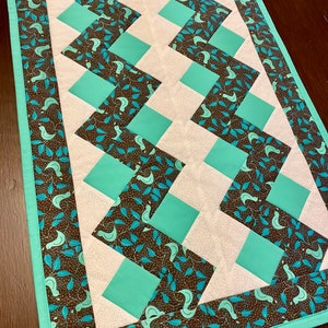 May include: A teal and brown quilted table runner with a chevron pattern. The runner has a border of brown fabric with a teal bird and leaf print.