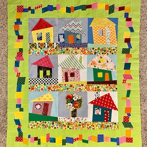 May include: A colourful patchwork quilt featuring a whimsical town of houses with different patterns and colours. The houses are surrounded by a green border with a colourful border of squares and rectangles.