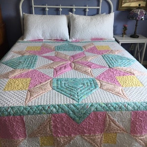 May include: A white bed with a white headboard and a white quilt with a pink, yellow, and green starburst pattern. The quilt is quilted with a variety of designs, including swirls, diamonds, and hexagons. The bed is made up with two white pillows. The headboard has a sign that reads "Count Your Blessings".
