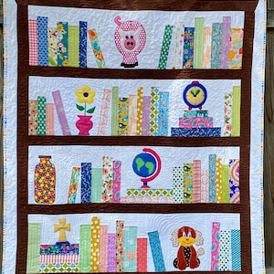 May include: A white quilted bookshelf with brown shelves and a white background. The shelves are filled with colourful books, and there are various decorative items on the shelves, including a pig, a flower, a clock, a globe, a cross, and a dog.