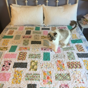 May include: A white bed with a white headboard and two white pillows. A white and gray cat is walking on a colorful patchwork quilt with a variety of patterns.