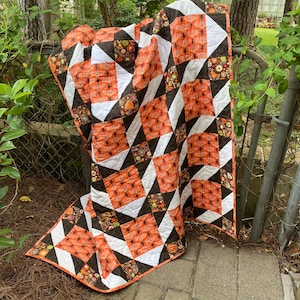 Op de afbeelding: Een quilt met een geometrisch patroon in oranje, zwart en wit. De quilt is gemaakt van stof met een bloemenprint en is gevouwen over een metalen hek.