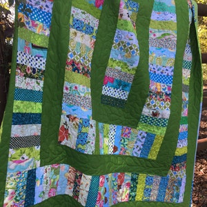 May include: A green patchwork quilt featuring a range of colourful fabrics in shades of blue, green, pink and white. The quilt is made up of strips of fabric sewn together to create a striped pattern. The quilt is hanging on a wooden fence.