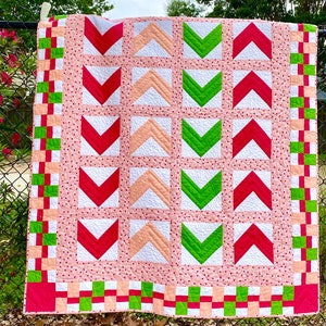 May include: A pink, white, and green quilt with a chevron pattern. The quilt is hanging on a fence.