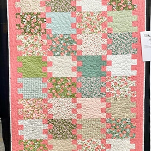 May include: A pink and green patchwork quilt with a floral pattern. The quilt is made up of squares and rectangles of different fabrics, all with a floral design. The quilt is quilted with a wavy pattern.