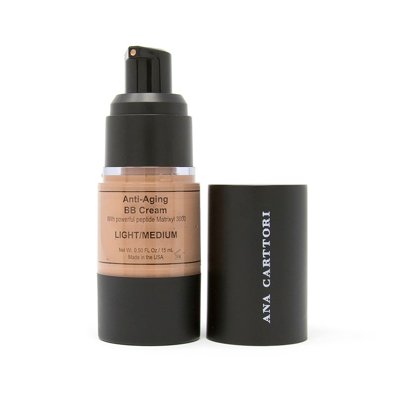 Op de afbeelding: Een zwarte en bruine pompflacon met anti-aging BB cr&egrave;me met de tekst "Anti-Aging BB Cream" en "LIGHT/MEDIUM" op het etiket. De flacon is gelabeld met "Net Wt. 0.50 FL Oz/15 ml" en "Made in the USA". De flacon ligt naast een zwarte dop met de tekst "ANA CARTTORI" erop gedrukt.