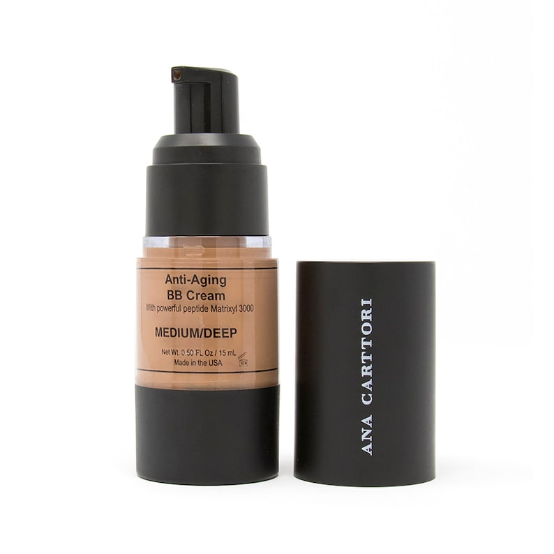 Op de afbeelding: Een zwarte en bruine fles anti-aging BB cr&egrave;me met de tekst "Anti-Aging BB Cream" en "MEDIUM/DEEP" op het etiket. De fles is ge&euml;tiketteerd met "ANA CARTTORI".