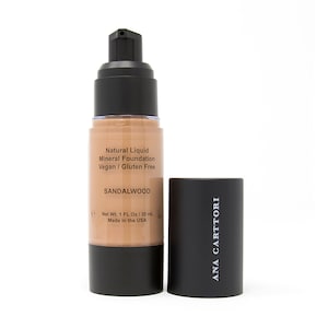 Op de afbeelding: Een zwarte en doorzichtige glazen fles met vloeibare minerale foundation met een zwarte pomp. De fles is gelabeld met "Natural Liquid Mineral Foundation Vegan / Gluten Free Sandalwood Net Wt. 1 FL Oz/30 mL Made in the USA" en "ANA CARTTORI".