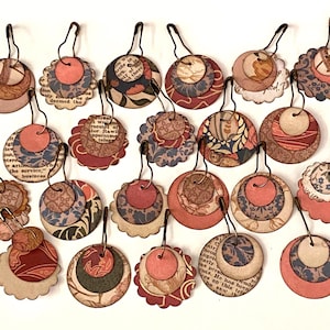 Puede incluir: Una colección de pendientes de papel hechos a mano con diseños circulares y festoneados en capas. Los pendientes presentan varios patrones, incluyendo florales, de texto y colores lisos en tonos rojos, azules y beige. Cada pendiente tiene un gancho de metal.