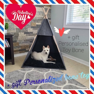 Pode incluir: Uma tenda tipi cinza e preta para cães com um cão branco dentro. A tenda está sobre um tapete cinza com um coração branco com asas e o texto "+ presente Osso de brinquedo personalizado".