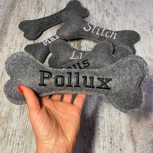 Op de afbeelding: Een set grijze vilten hondenbot speelgoed met geborduurde namen, waaronder "Max", "Stitch", "Lil' Gus" en "Pollux".