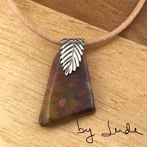 May include: A brown, green, and red stone pendant with a silver leaf charm on a brown leather cord. The pendant is irregularly shaped and has a mottled appearance. The text "by Jude" is written on the wooden surface below the pendant.