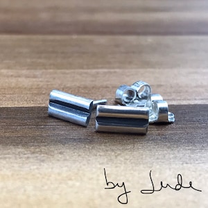 May include: A pair of silver earrings with a rectangular shape and a grooved design. The earrings are on a wooden surface with the text "by Jude" written below.