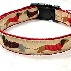 May include: A red and beige dog collar with a black plastic buckle. The collar features a repeating pattern of dachshund dogs wearing red sweaters.