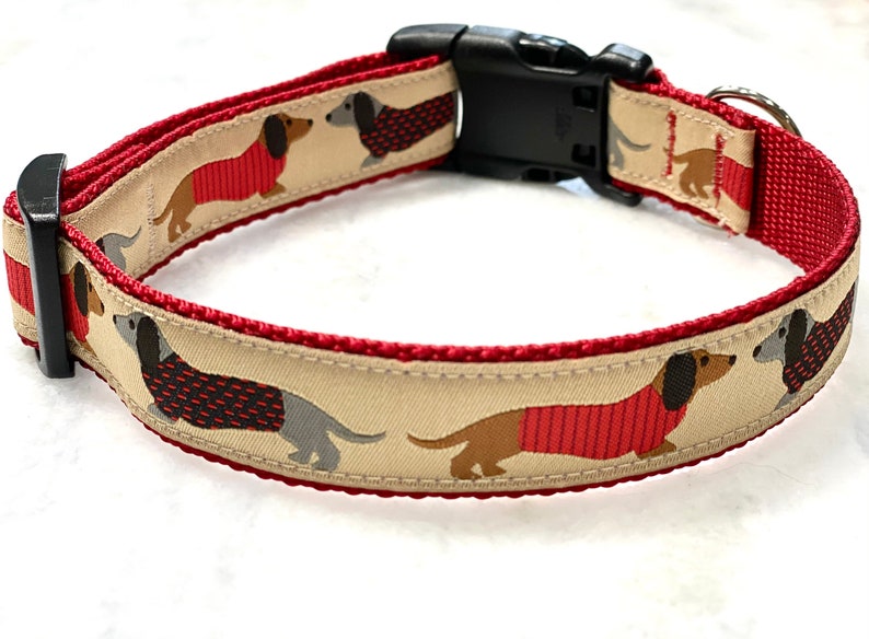 May include: A red and beige dog collar with a black plastic buckle. The collar features a repeating pattern of dachshund dogs wearing red sweaters.