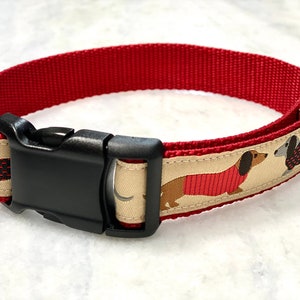 May include: Red dog collar with a beige and red dachshund pattern. The collar has a black plastic buckle and a silver D-ring.